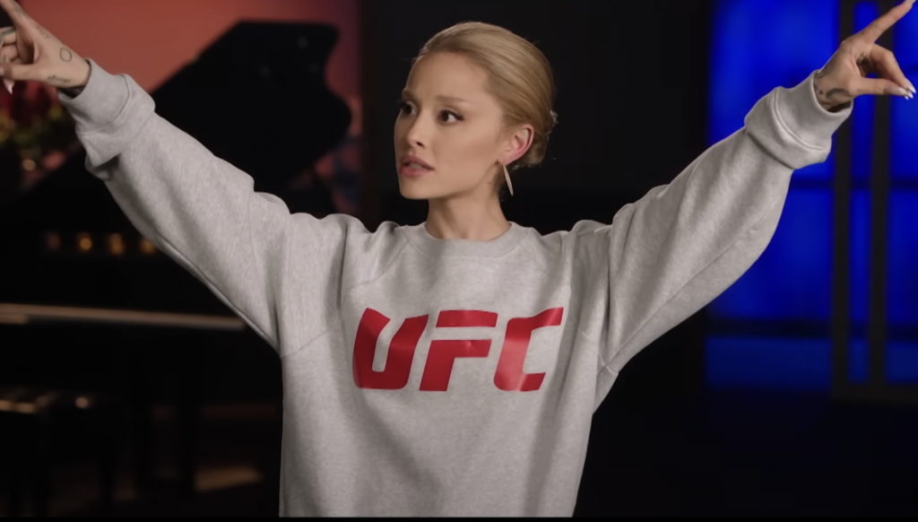 Ariana Grande impersonating Celine Dion wearing a UFC sweater with arms raised and pointing in opposite directions