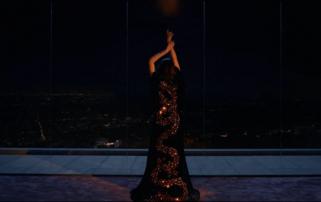 Demi Moore in The substance in a long sparkly gown in front of a floor to ceiling window with arms up