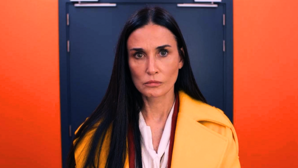 Demi Moore in 2024 The Substance looking in a mirror in a yellow jacket with a red wall, blue door in the background