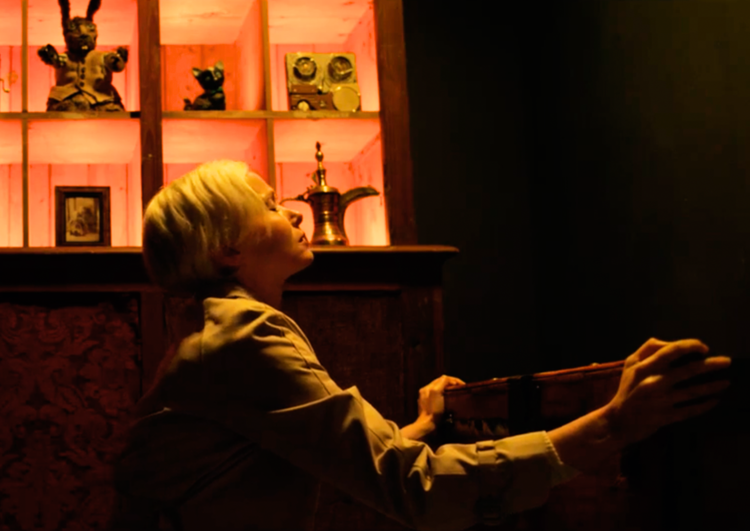 Scene from The Oddity where Dani is in a dark room opening a chest with her eyes closed, glowing orange lighting behind her