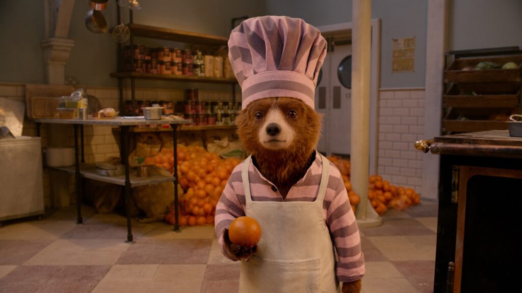 Paddington in a pink and grey prison uniform holding an orange.