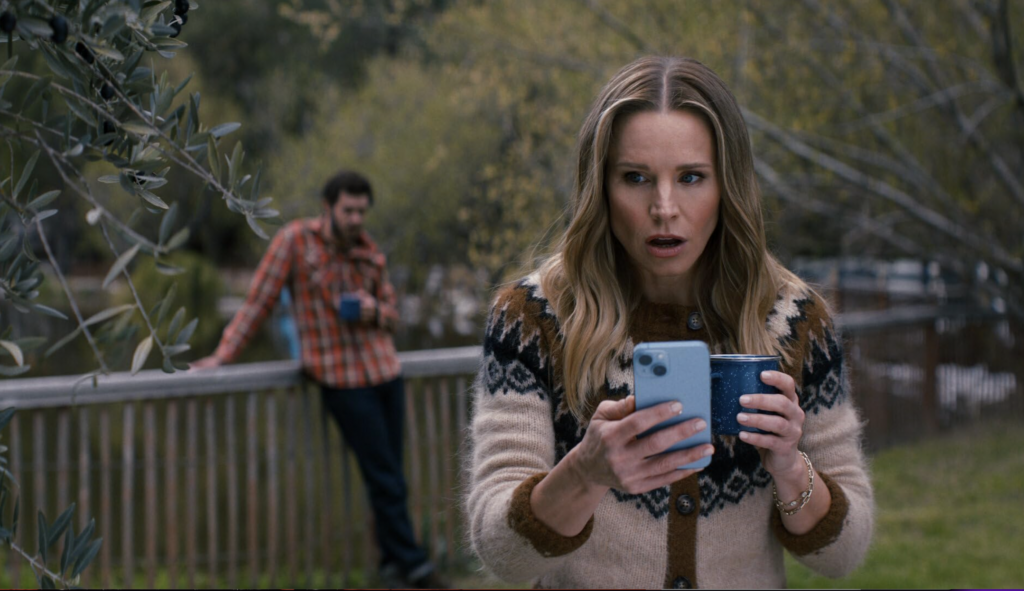 Kristin Bell looks at her phone with a surprised face holding a cup of coffee. A blurry Adam Brody can been see in the outdoorsy background, a scene from Nobody Wants this