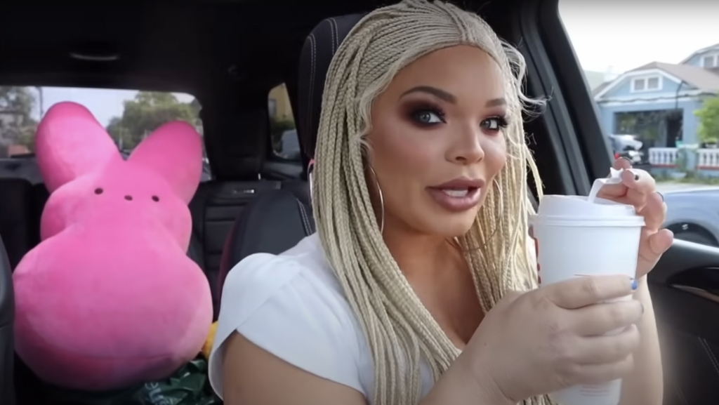 Trisha Paytas in her car wearing cornrows, drinking a coffee with a big pink Peep plushie in the background