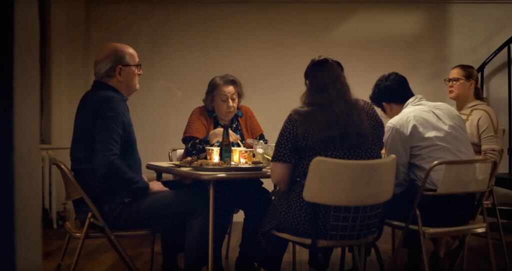 A family sits around a fold out table on fold out chairs in a run down apartment in 'The Humans.'